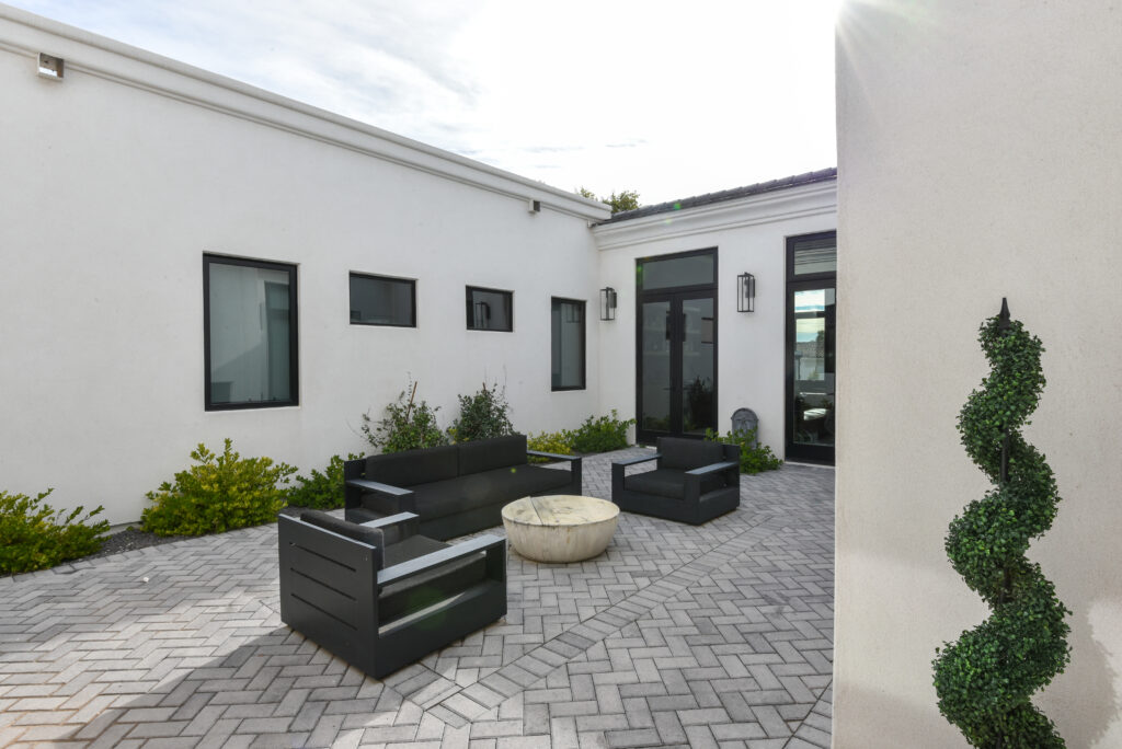 Enclosed outdoor courtyard framed by white stucco walls and black-trimmed windows, furnished with a modern black outdoor sofa and matching armchairs around a round stone coffee table on gray herringbone brick pavers in bright daylight