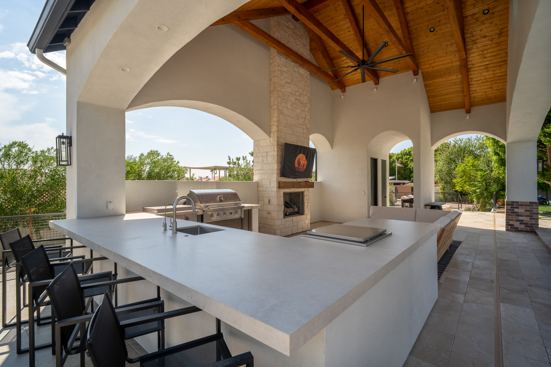 Outdoor kitchen in the cabana with a large white bar island and dark contemporary barstools, a stainless-steel prep sink and a built-in cooktop under the vaulted wood ceiling, the stacked-stone fireplace beyond