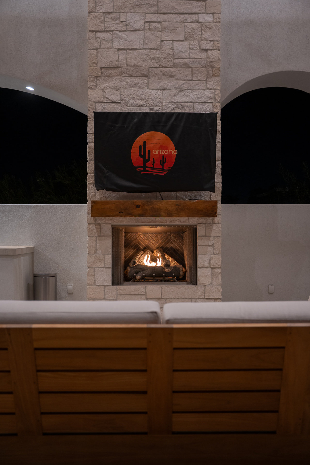 Night view of the covered patio with a light stacked-stone fireplace and rustic wood mantel, an active fire and a covered television above, the slatted back of a natural wood sofa with off-white cushions in the foreground