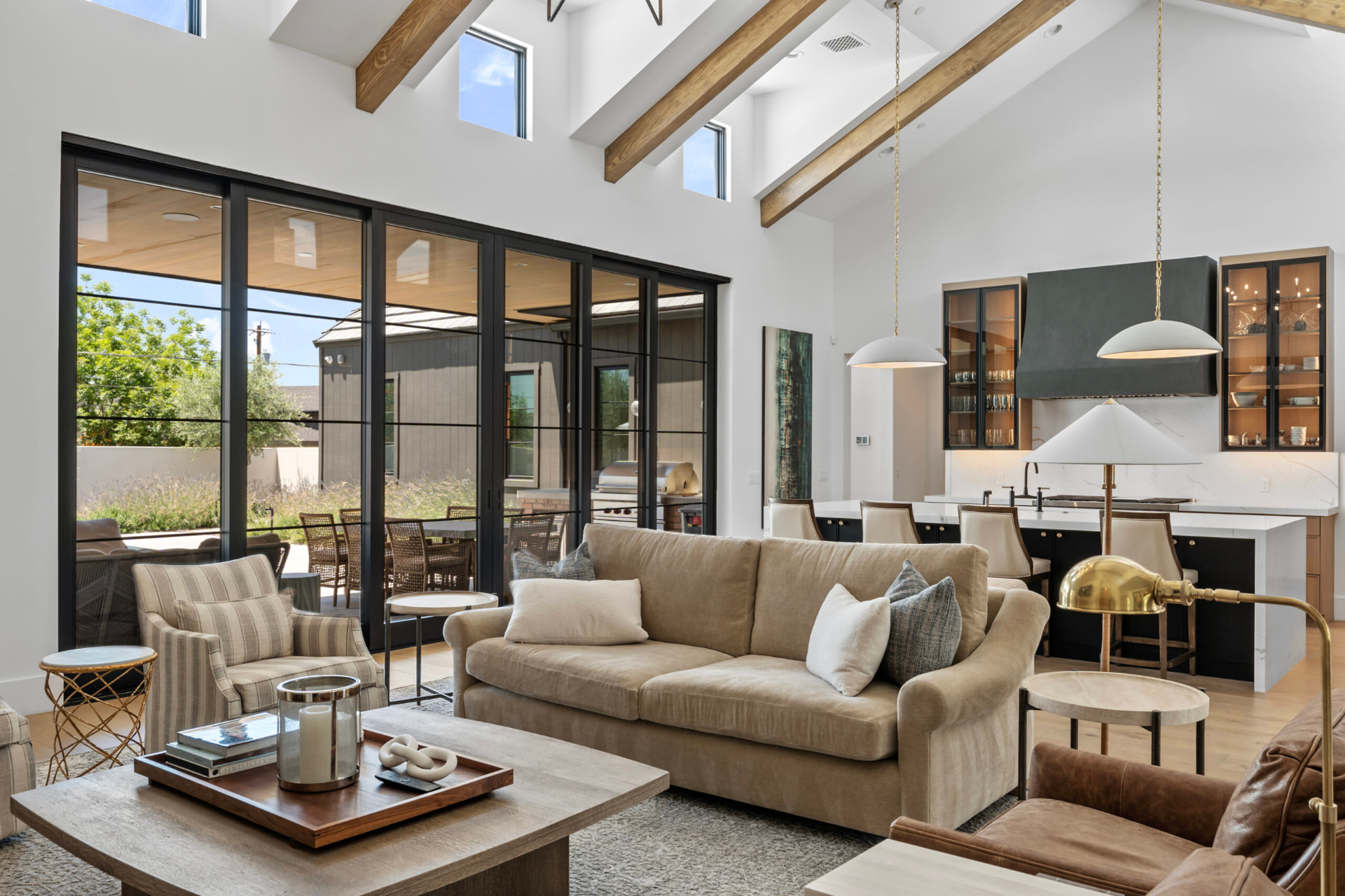 Living room with a vaulted beam-filled ceiling and black-framed sliding glass doors opening to the backyard patio, furnished with a neutral beige sofa, accent armchairs and a wood coffee table, flowing into the kitchen with a dark island, marble counters and modern pendant lights