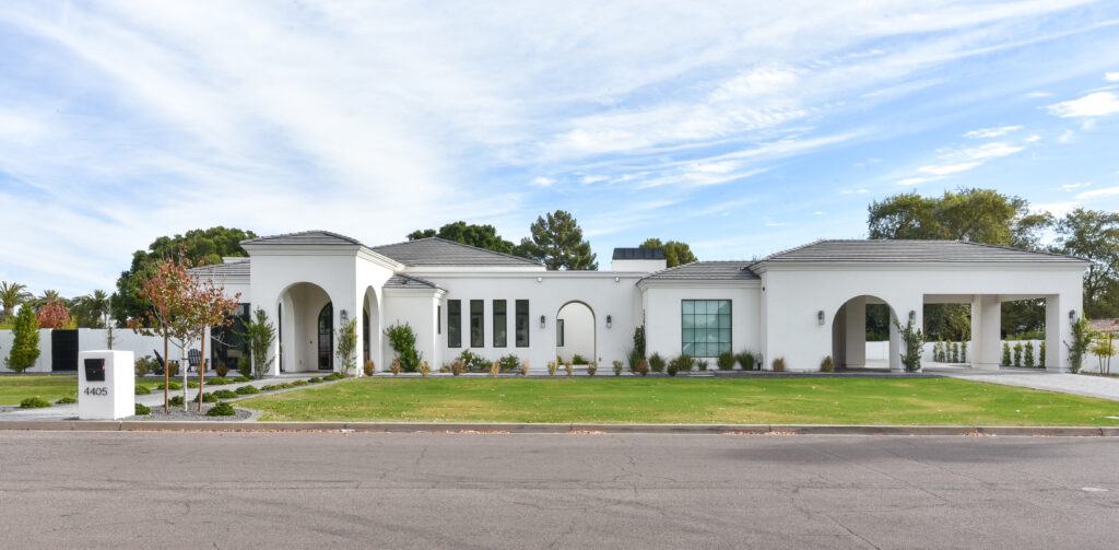 Straight-on street view of the single-story modern Mediterranean home with crisp white stucco walls, dark gray concrete roof tiles, arched entryways and black-framed grid windows, across a manicured lawn with a white pillar mailbox under a bright blue sky