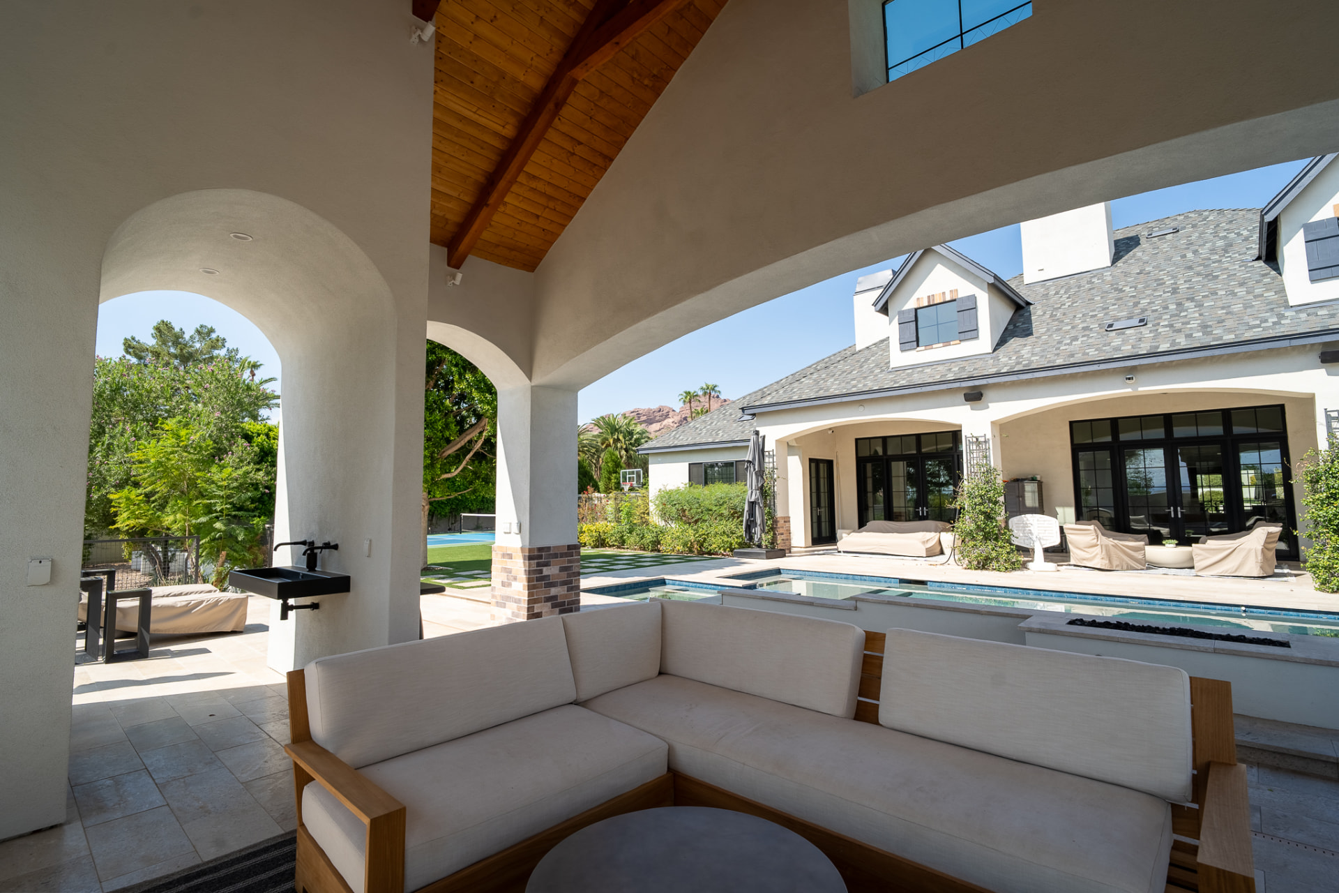 Shaded poolside pavilion with a light-cushioned wooden sectional sofa and a wall-mounted black exterior sink beneath an exposed wood beam ceiling, the wide arched opening framing the turquoise pool and the main white stucco residence beyond