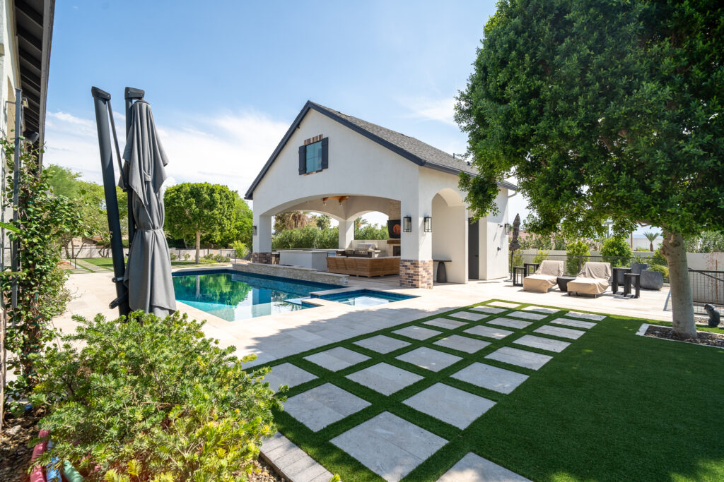 Backyard with a rectangular swimming pool and attached hot tub beside an open-air white stucco pool cabana with arched openings, light stone pavers set in a turf grid, lounge chairs and a closed patio umbrella