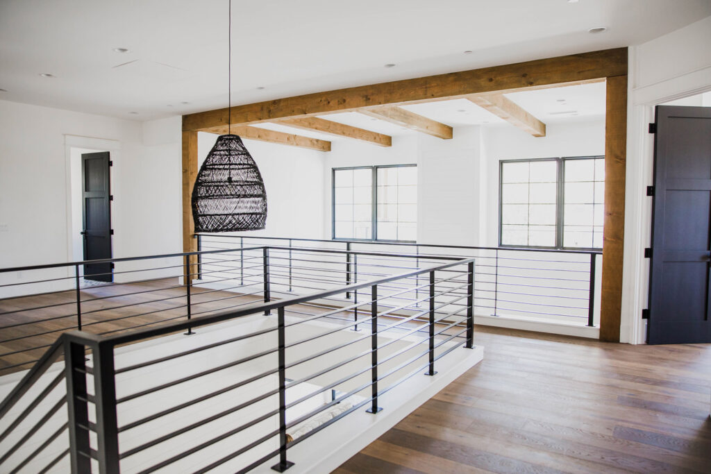 Second-story landing with hardwood floors, rustic ceiling beams, black railings around the open stairwell and black-trimmed grid windows