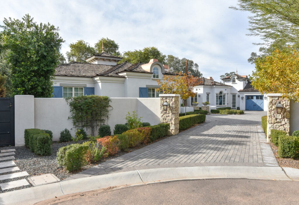 Long gray paver driveway flanked by white privacy walls and natural stone pillars with lanterns, leading to a one-story residence with white stucco walls, weathered wood shake roofing, dormer windows and powder blue shutters and garage doors, framed by boxwood hedges