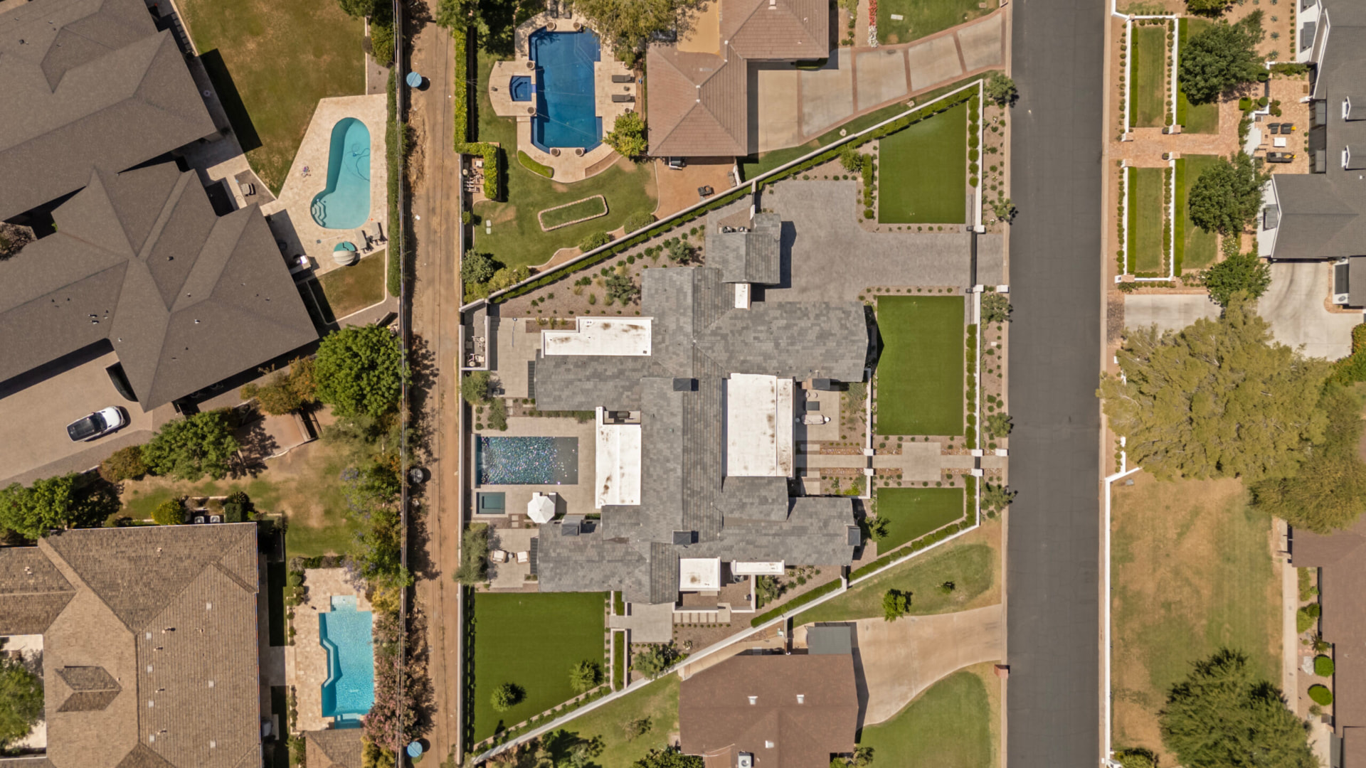 Overhead aerial view of the property showing the sprawling dark-gray tiled roof, the geometric backyard pool, surrounding sun patios and manicured turf panels, with neighbouring streets and yards beyond