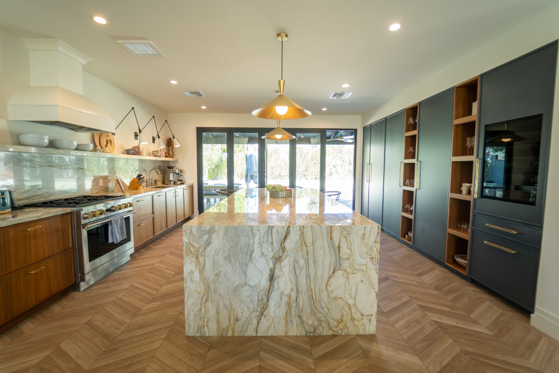 Kitchen with a large marble waterfall island, wood lower cabinets, a stainless steel range, brass cone pendants and charcoal full-height cabinets with open wood shelving