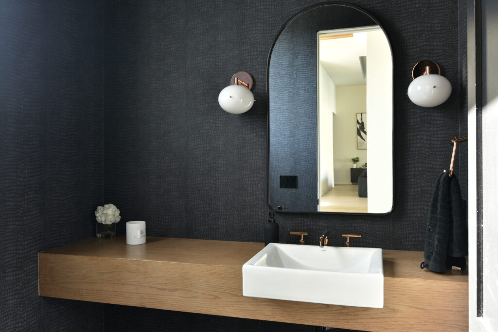 Powder room with dark charcoal textured wallpaper, a floating natural wood vanity and a white rectangular vessel sink with warm metal hardware, beneath an arched black-framed mirror flanked by two round frosted globe sconces