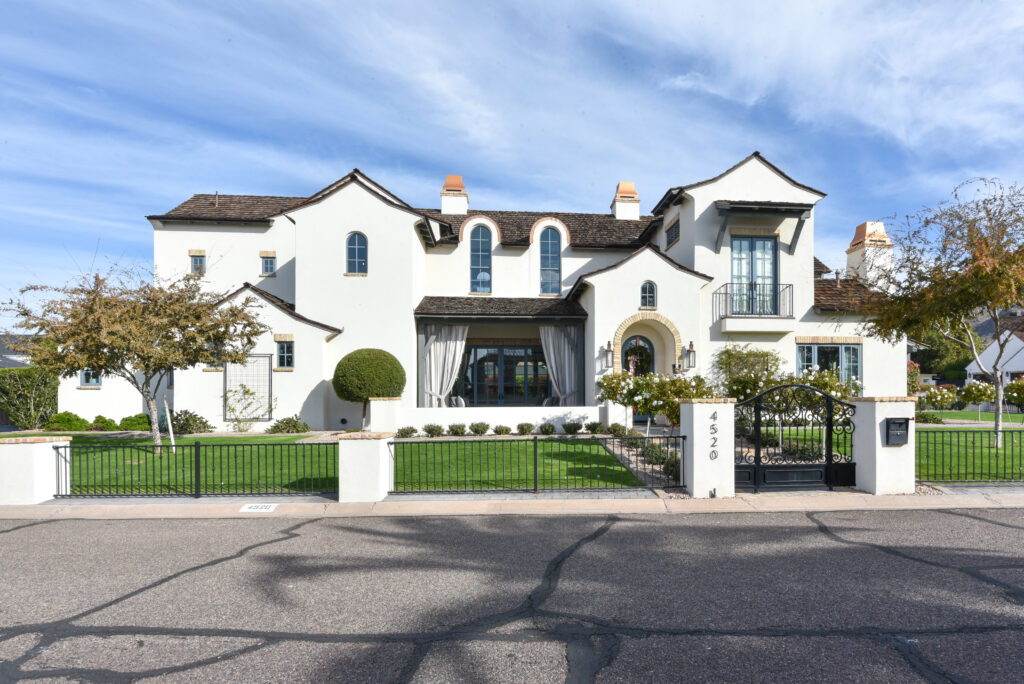 Front exterior with multi-gabled dark rooflines, terracotta-accented chimneys and an arched stone entryway, a covered front porch with outdoor drapery and a decorative black iron gate along the street