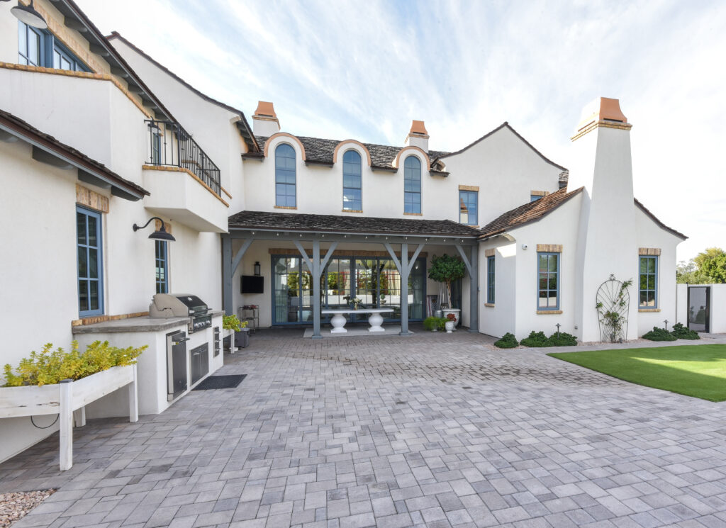Backyard patio with grey brick pavers, a built-in outdoor kitchen with a stainless steel grill and a covered porch on slate-grey posts, beneath the multi-story white stucco facade with slate-blue trimmed arched windows and a second-floor balcony
