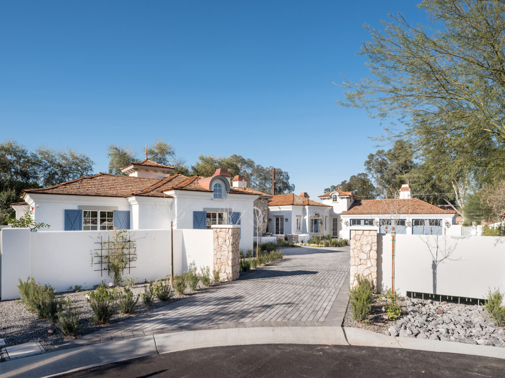 Stone-paved driveway flanked by white boundary walls, stacked stone pillars and low gravel garden beds, with the single-story white stucco estate beyond showing terracotta tile roofs, dormer windows and light blue shutters and garage doors