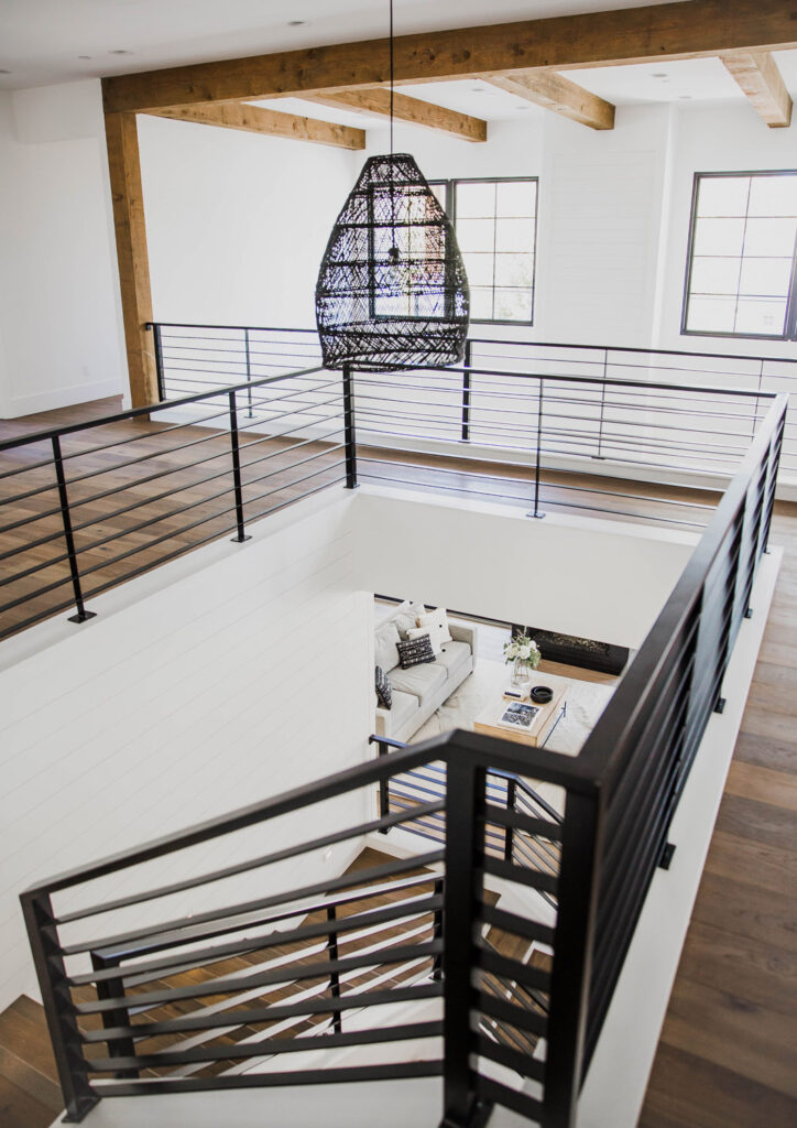 View from the upper landing down the stairs with black metal railings into the living room with a light grey sofa beneath exposed timber beams
