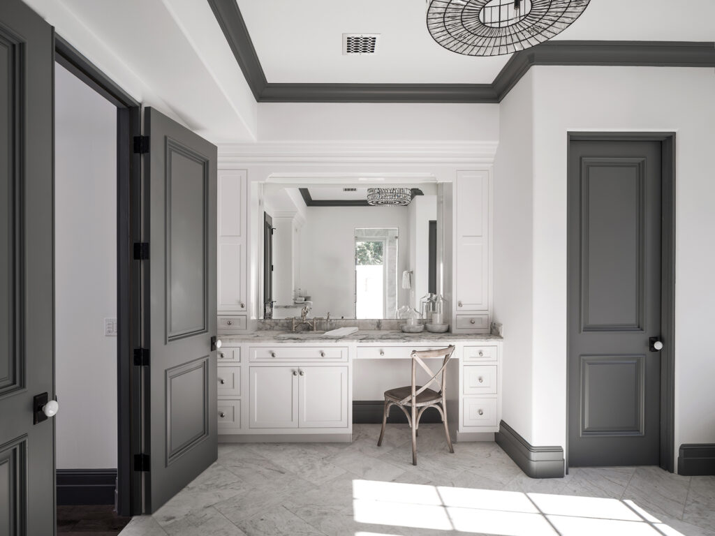 Bathroom with a custom white makeup vanity and sink station with marble countertops, a large framed mirror, a wooden cross-back chair and dark charcoal paneled doors and trim