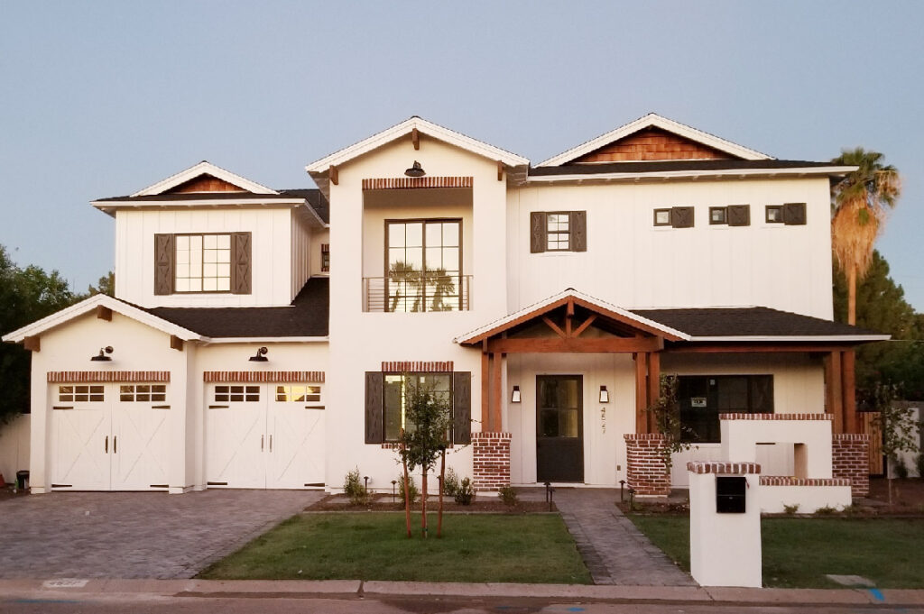Exterior of a two-story modern farmhouse with white siding, dark-framed windows, red brick pedestals, a wood-timbered front porch and carriage-style garage doors along a paved driveway