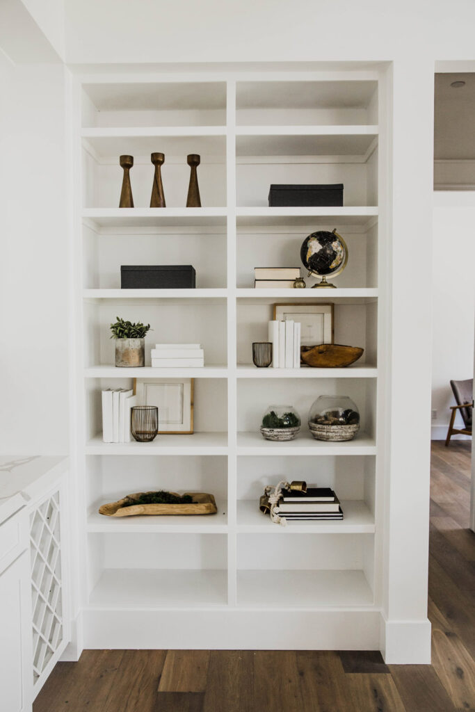 Built-in white shelving with brass candlesticks, books, a globe and decorative accents beside a wine rack cabinet above light wood floors