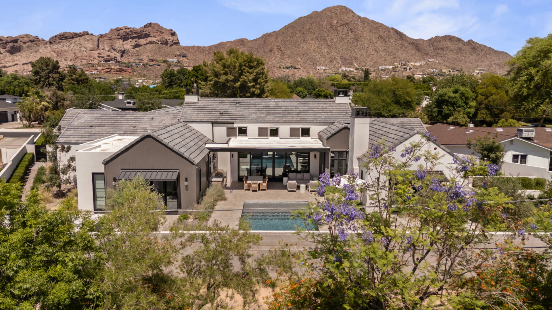 Elevated view of the rear exterior and patio against Camelback Mountain, with a rectangular inground swimming pool, a stone patio set with outdoor dining and lounge furniture, a covered pergola and flowering jacaranda trees framing the property