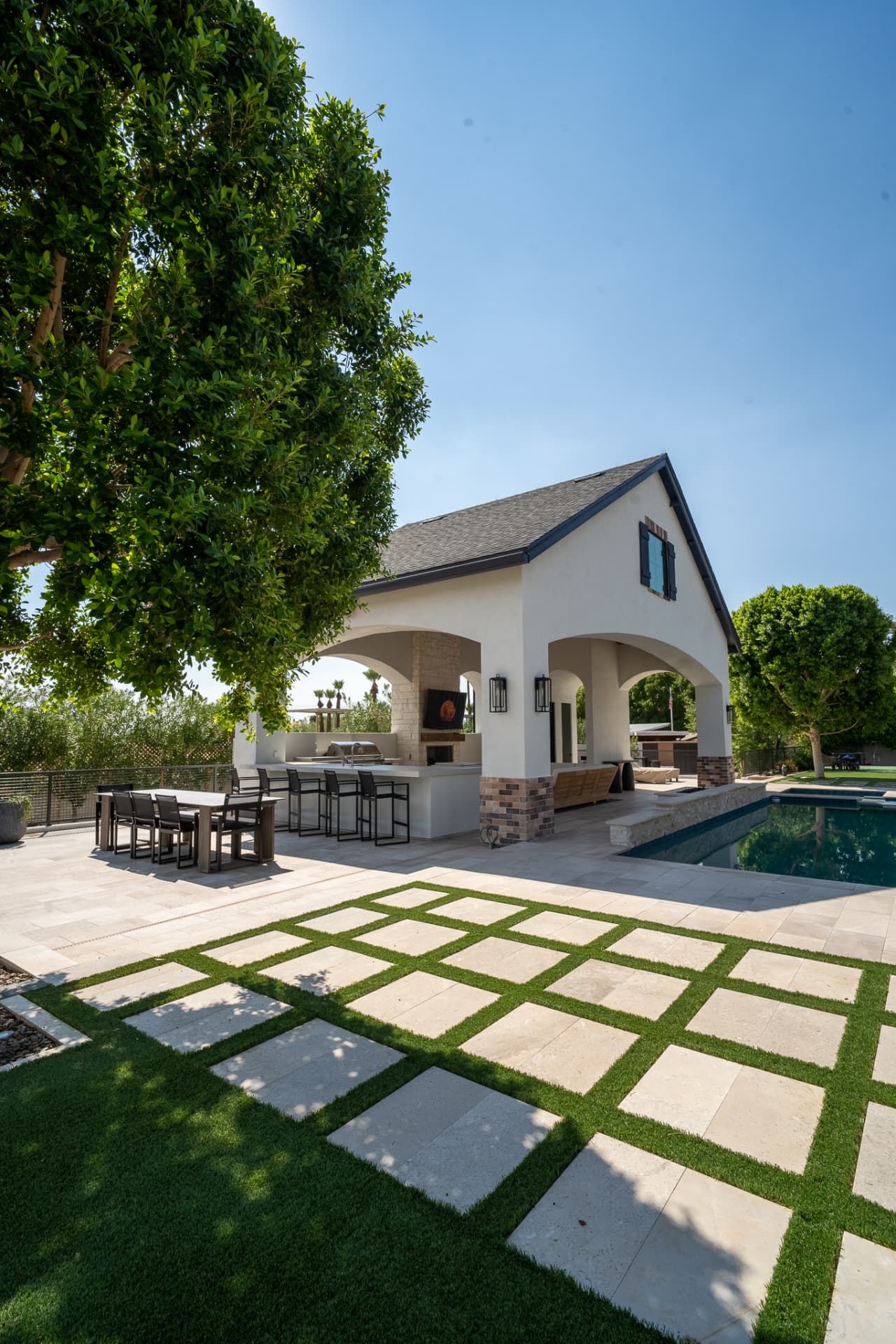 Outdoor kitchen and dining area under the pavilion with a bar counter and stools beside an outdoor dining table, light stone pavers interspersed with green turf in a geometric grid next to the swimming pool
