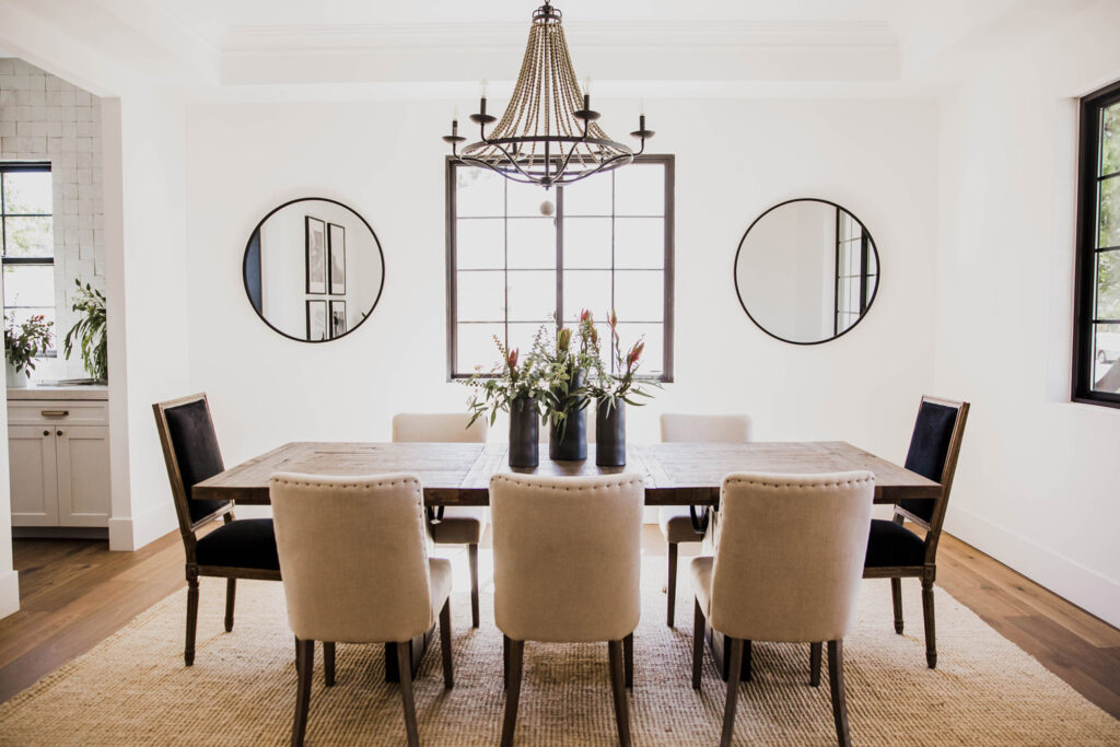 Dining room with a rustic wood table, beige and black upholstered chairs, a beaded iron chandelier and two round black-framed mirrors flanking a central window