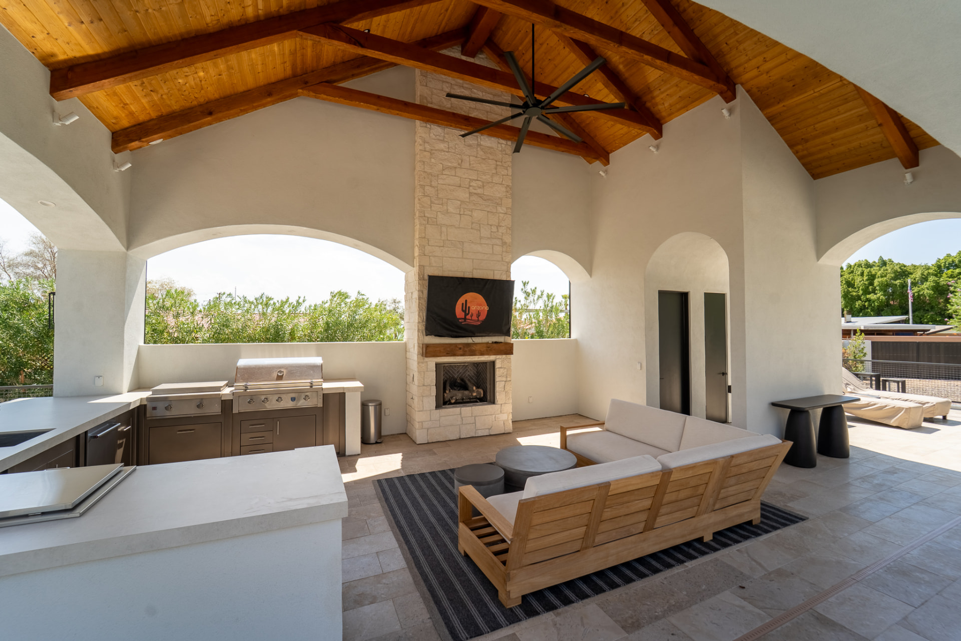 Inside the open-air poolside cabana with a vaulted exposed-wood beam ceiling and a black ceiling fan, a wooden sectional sofa on a striped rug facing a stacked-stone fireplace with a mounted television and a stainless-steel outdoor kitchen counter