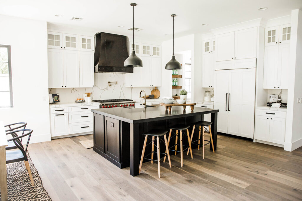 Kitchen with white shaker cabinetry, a large black island with three barstools, black dome pendants, a dark range hood and marble-look countertops on light oak floors
