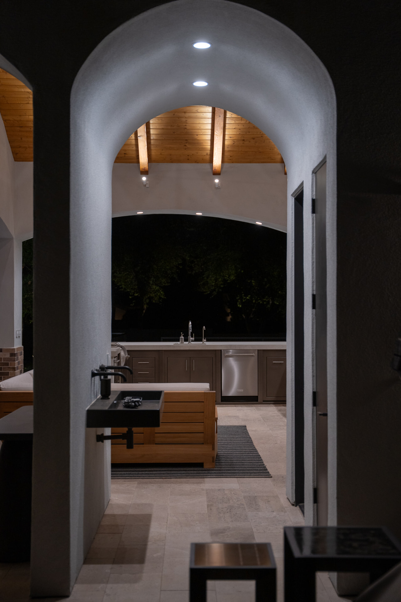 Night view through an arched white stucco breezeway into the pool cabana, where recessed lights illuminate the vaulted wood-slat ceiling, a black wall-hung sink, brown cabinetry with stainless-steel appliances and a wooden outdoor sofa