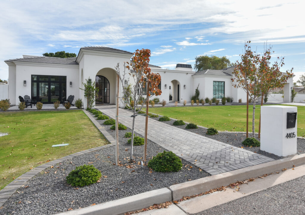 Gray herringbone paver walkway leading past dark gravel beds, young saplings and shrubbery to the home's arched front entrance, with a front patio and dark Adirondack chairs to one side and a white stucco mailbox pillar at the sidewalk