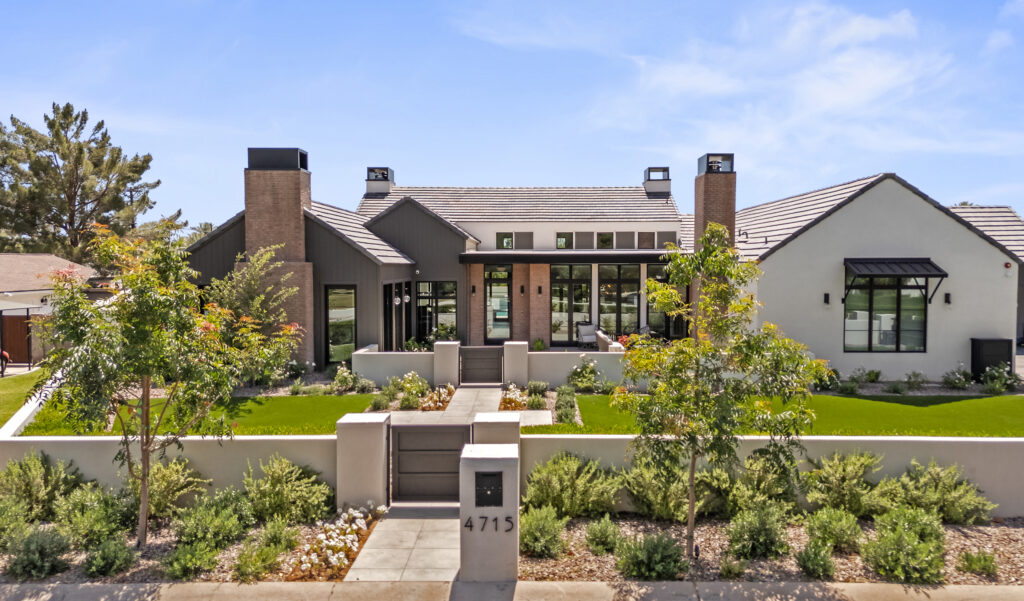 Elevated view of the front exterior with white stucco walls, dark charcoal vertical siding, brick chimneys and dark-framed picture windows, behind a low perimeter stucco wall and entry gate framing a manicured lawn, garden beds and a stone walkway