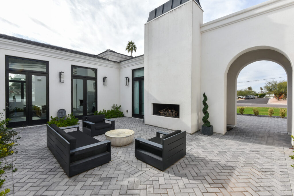 Wide view of the paved courtyard patio showing the white stucco fireplace chimney, the black outdoor sofa set with a round stone table, and tall black-framed French doors, with an arched opening looking out toward the neighborhood street