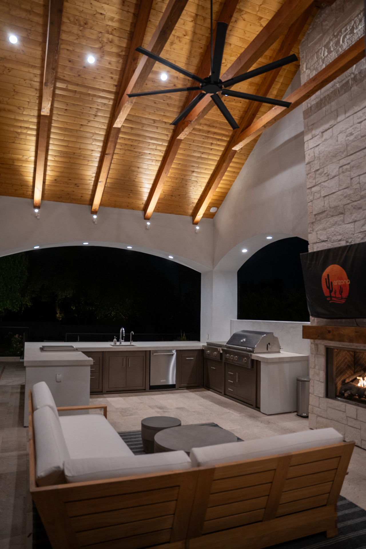 Night view of the covered patio and kitchen pavilion beneath a vaulted tongue-and-groove wood ceiling with exposed beams and warm recessed lighting, an L-shaped outdoor kitchen with brown cabinetry and stainless-steel appliances, and a wood-framed sectional sofa facing the stone fireplace column