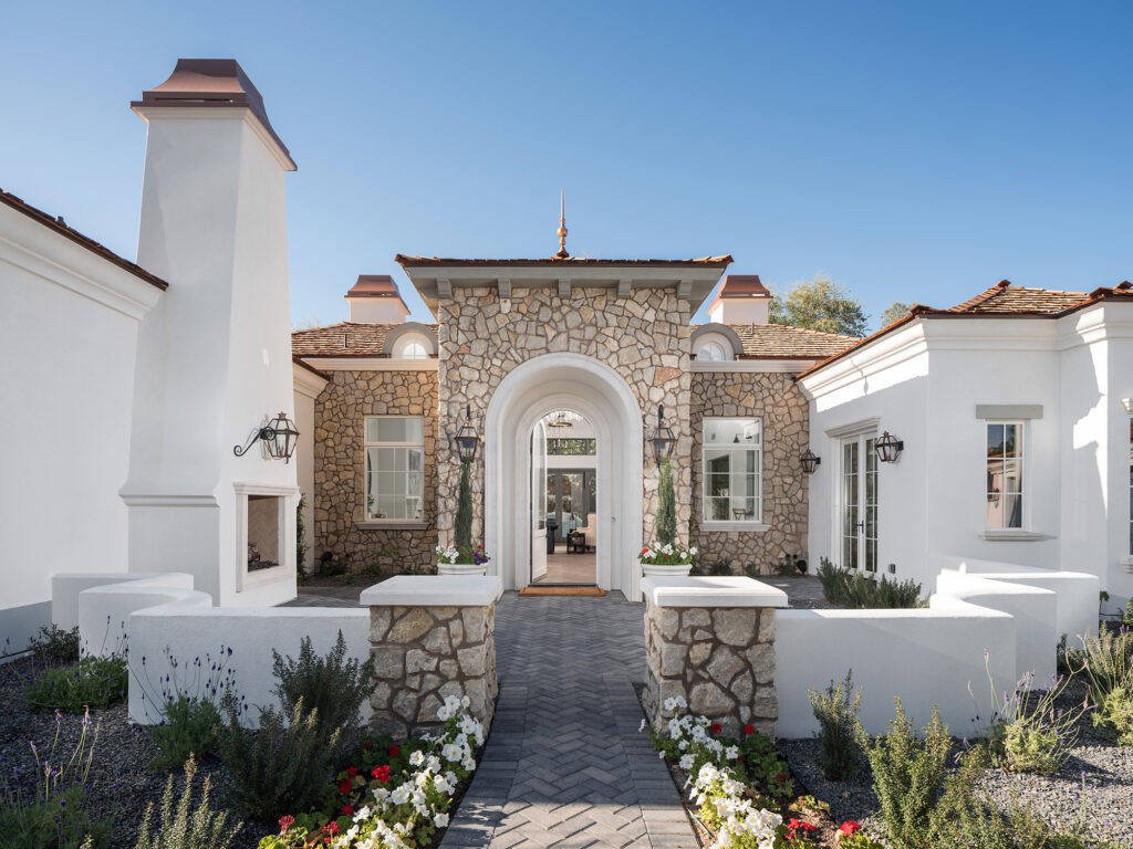 Front entrance with a central stone-clad arched doorway, white stucco wings with a tall outdoor fireplace, wrought-iron wall lanterns and a herringbone paver path flanked by stone pillars and blooming flowerbeds