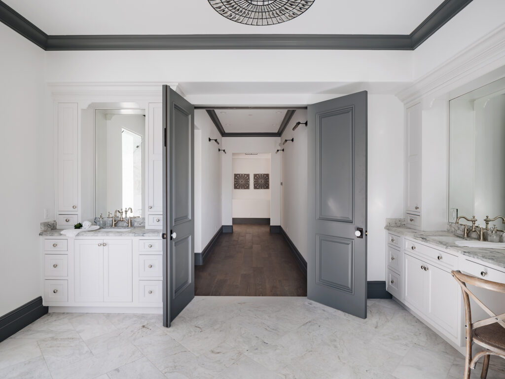 Bathroom with twin white vanities and gray-veined marble countertops, brass faucets and large mirrors against white walls with charcoal crown molding, open charcoal double doors leading to a hallway with dark hardwood floors