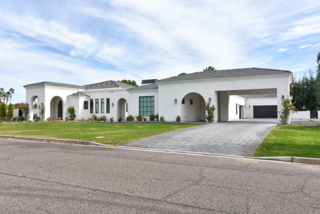 Angled front exterior of the white stucco home with a gray herringbone paver driveway running under an integrated porte-cochère toward a black garage door, with tidy desert shrubs, a green front yard and black wall sconces in clear daylight