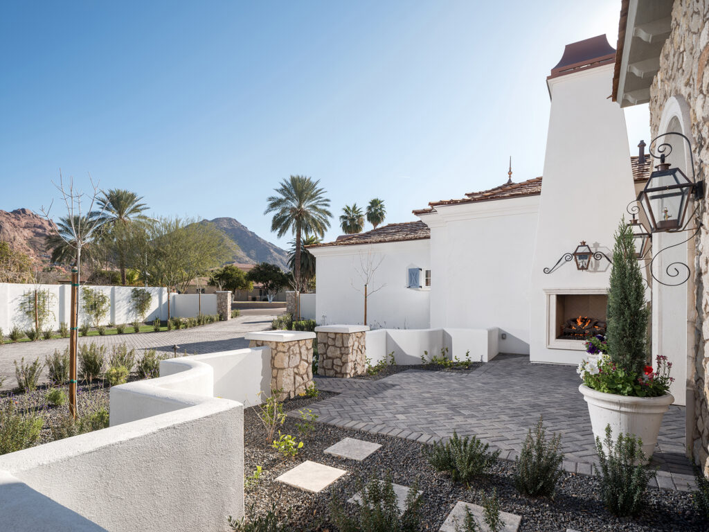 Private courtyard with a gray paver patio, a tall white stucco outdoor fireplace, stone pillars and low curved white walls above a paved driveway with palm trees and desert mountains beyond