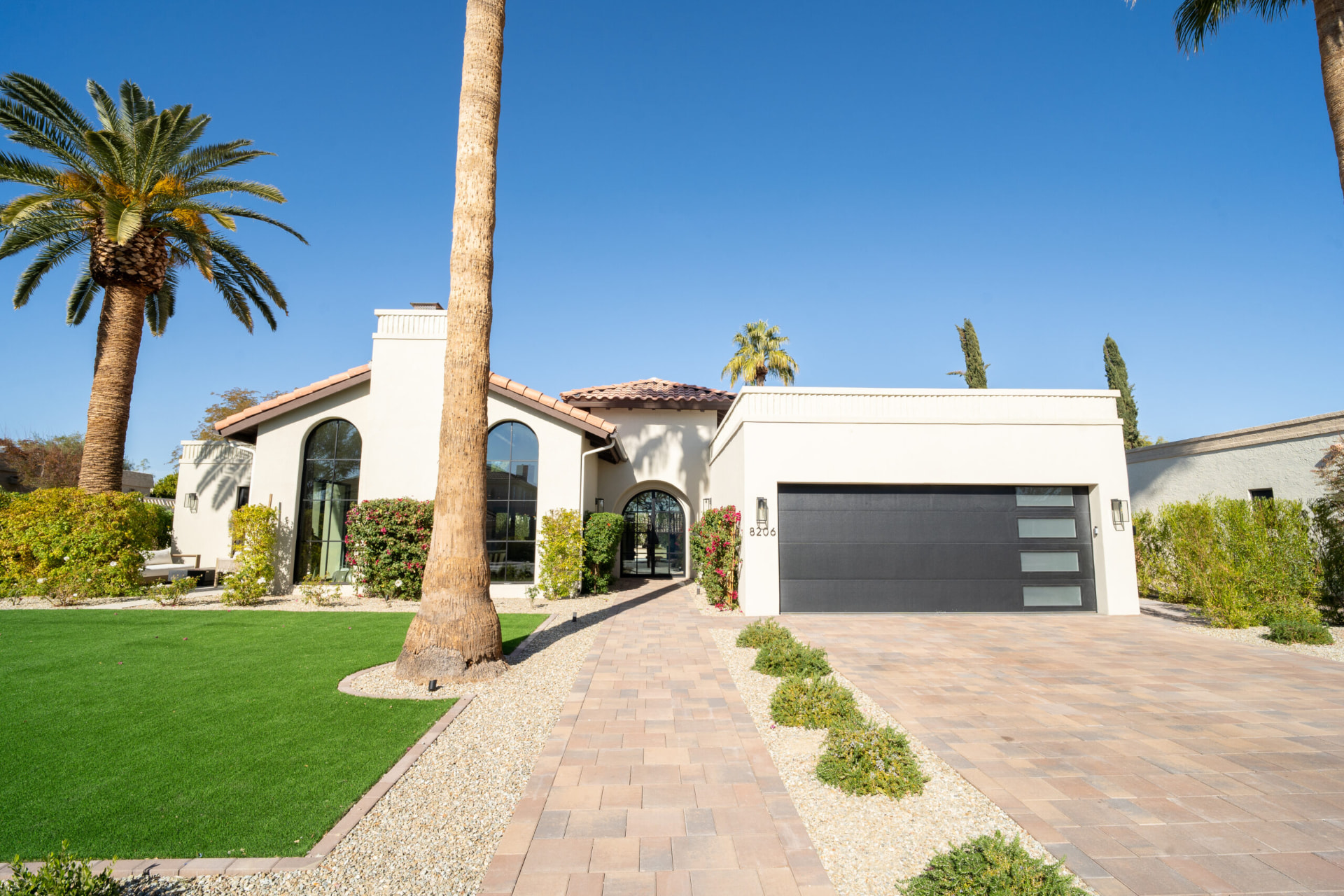 Front exterior of a single-story cream stucco home with terracotta roof accents, arched black-framed windows, a dark garage door, paver walkways and desert landscaping with palm trees