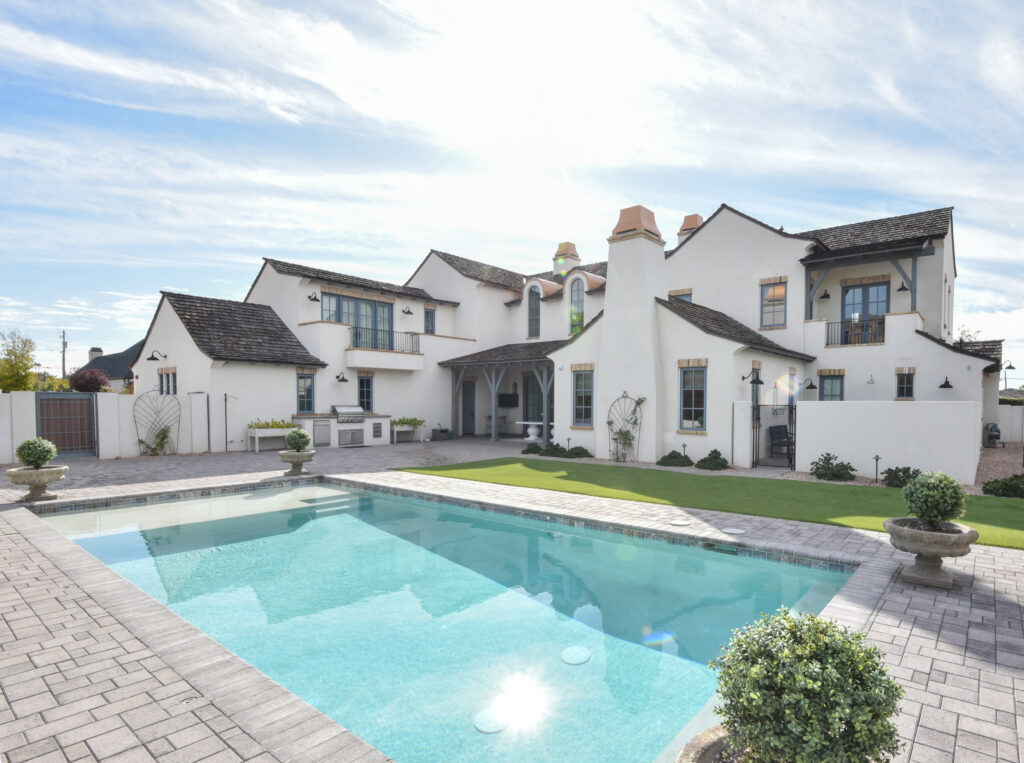 Backyard swimming pool with turquoise water surrounded by grey stone pavers, topiary urn planters and a green lawn, with the full rear facade of the white stucco estate showing dark shake-shingle gables, slate-blue window trims and a covered terrace
