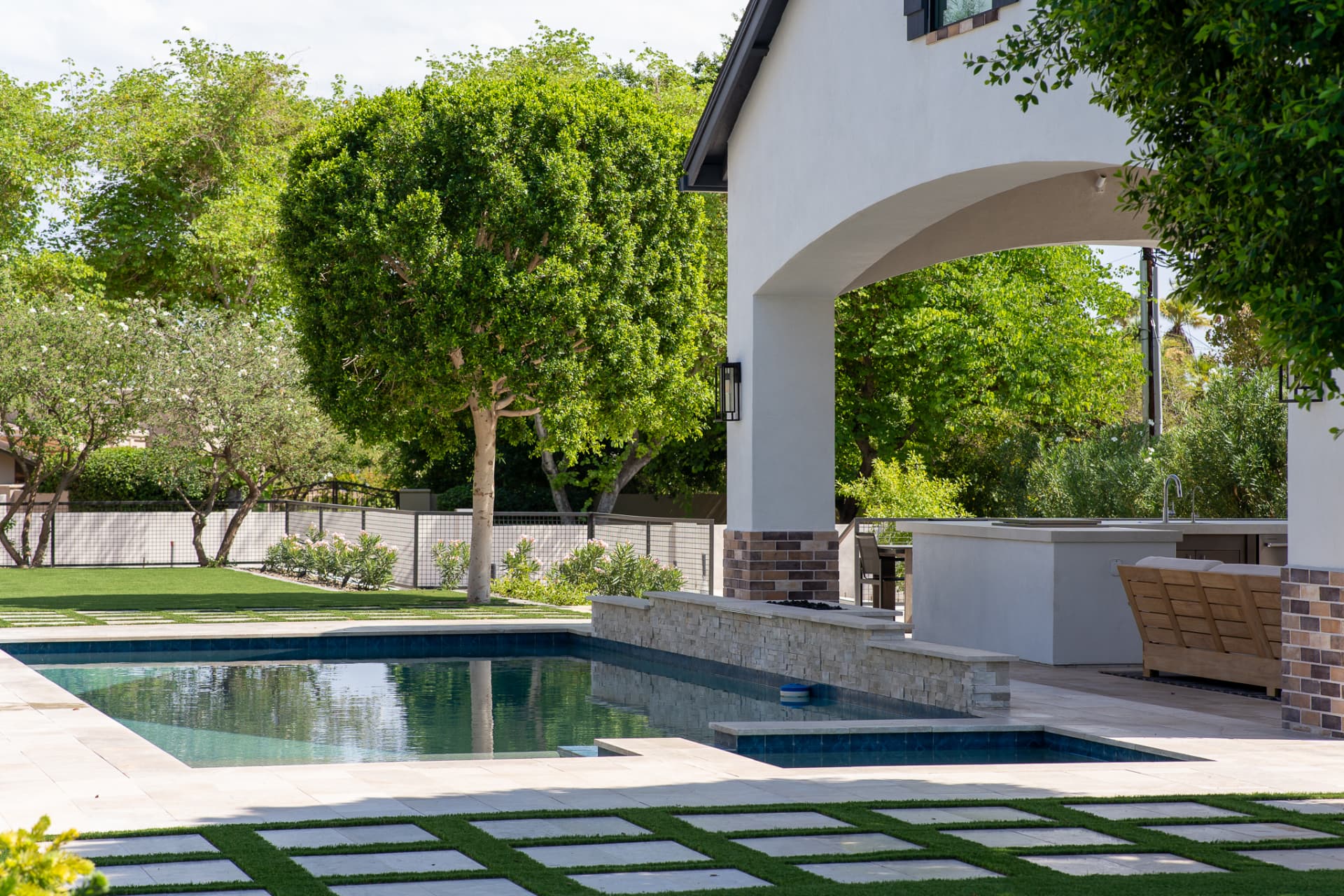 Swimming pool and hot tub bordered by a raised stacked-stone water wall beside the white arched pool cabana, with square light-gray stone pavers set in artificial turf and green trees beyond