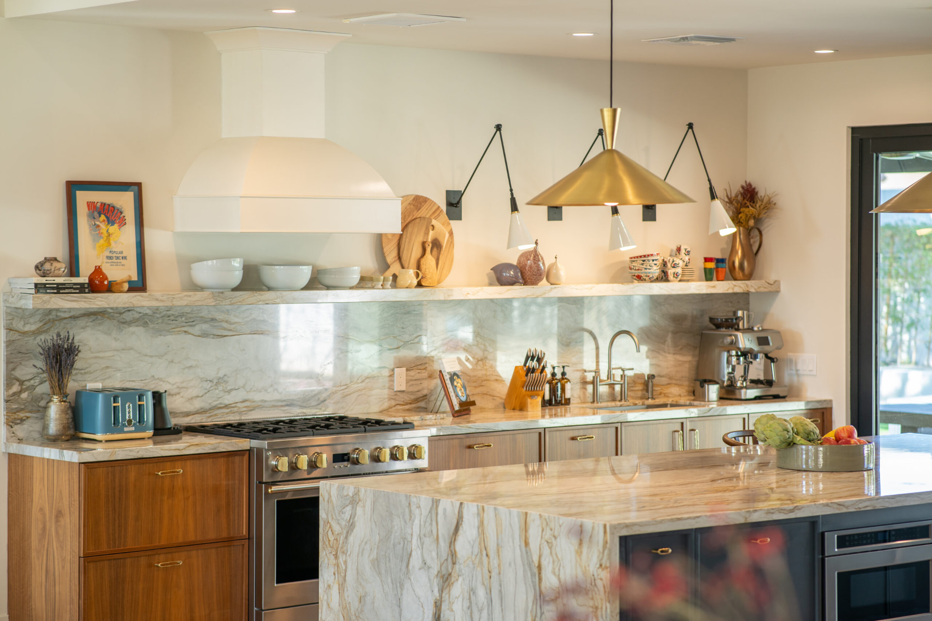 Kitchen with a marble waterfall island and matching slab backsplash, warm wood lower cabinets, a white range hood over a stainless gas range and brass cone pendants