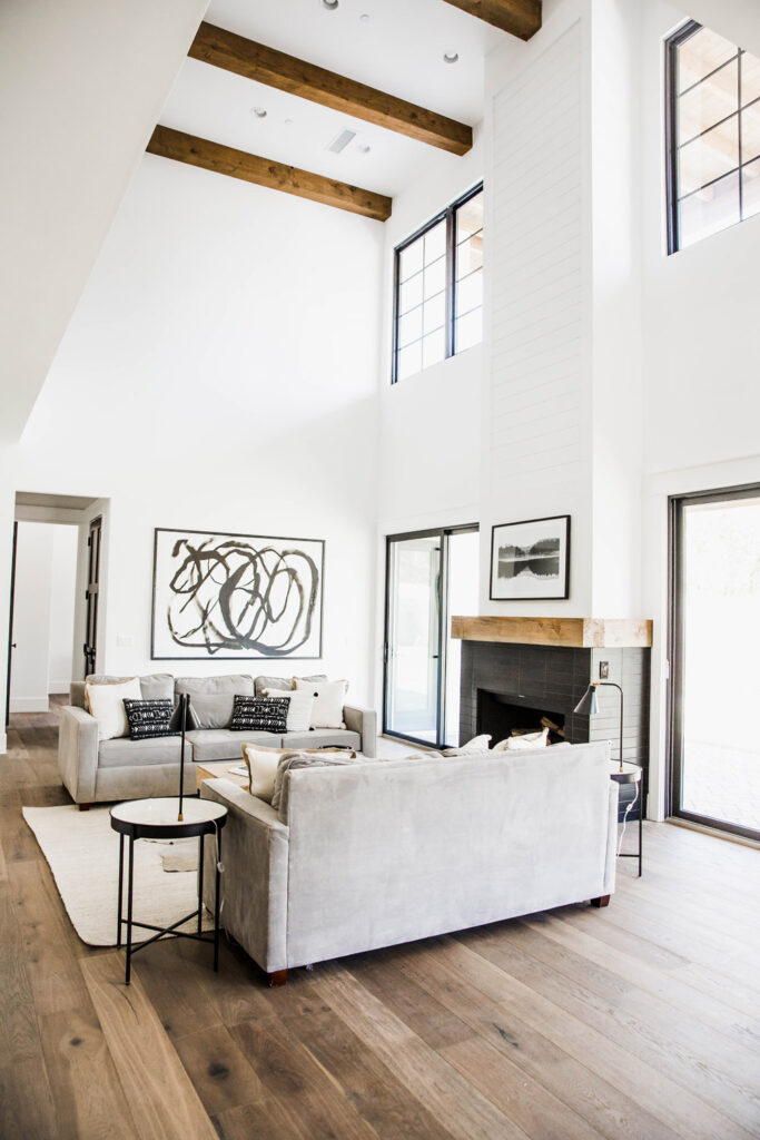 Living room with high ceilings, exposed timber beams and clerestory windows, grey sofas around an area rug and a dark tile fireplace topped by a wood mantel beam