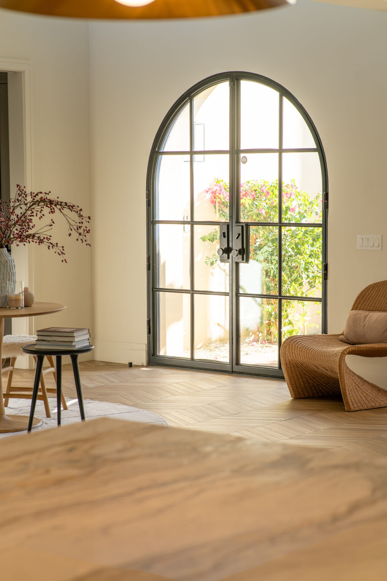 Room with light oak herringbone floors, a sculpted wicker lounge chair and a round wooden table beside a black-framed arched glass door looking out to a sunlit courtyard