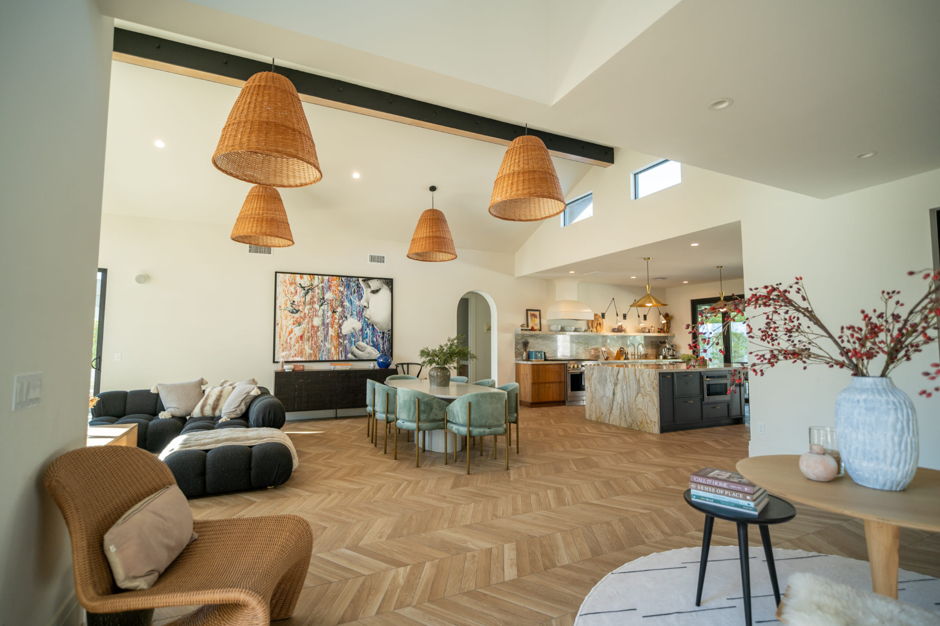 Open great room with vaulted ceilings, an exposed dark beam, woven pendant lights, a dark tufted sectional and a dining table with light blue-green velvet chairs