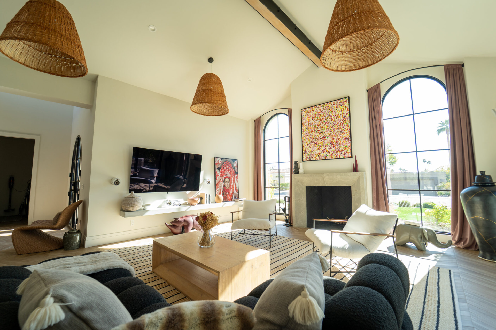 Living room with a stone fireplace, tall arched black-frame windows, woven pendant lights and dark curved seating around a light wood coffee table