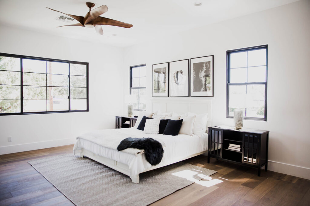 Bedroom with a white bed, dark nightstands and black-trimmed windows beneath three black-and-white art prints and a wooden ceiling fan