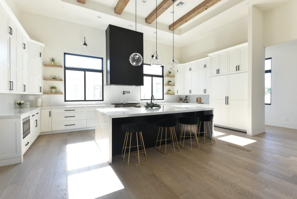 Bright kitchen with white shaker cabinetry, a black herringbone range hood, a white waterfall island with a black base and gold-legged black stools, clear globe pendants and exposed wood ceiling beams