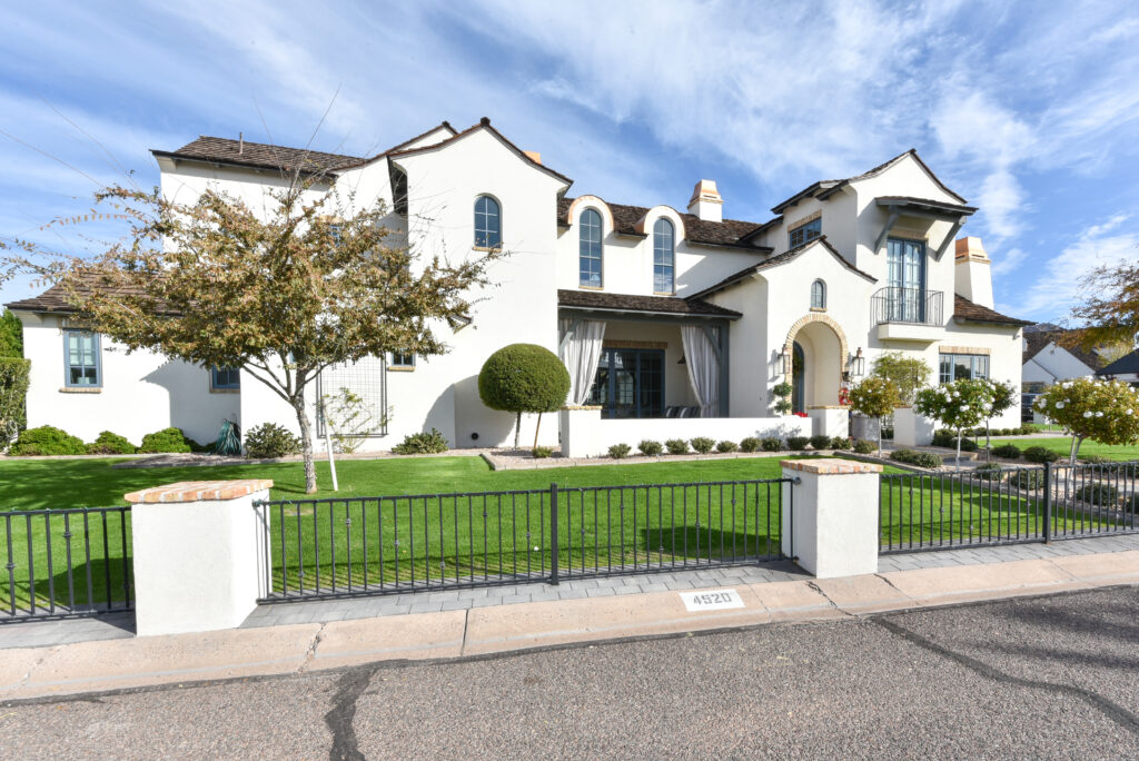 Angled exterior view of the white stucco residence showing a black metal fence with brick-topped white pillars along the curb, a covered veranda with sheer white drapes, rounded topiary bushes and an upper-level balcony