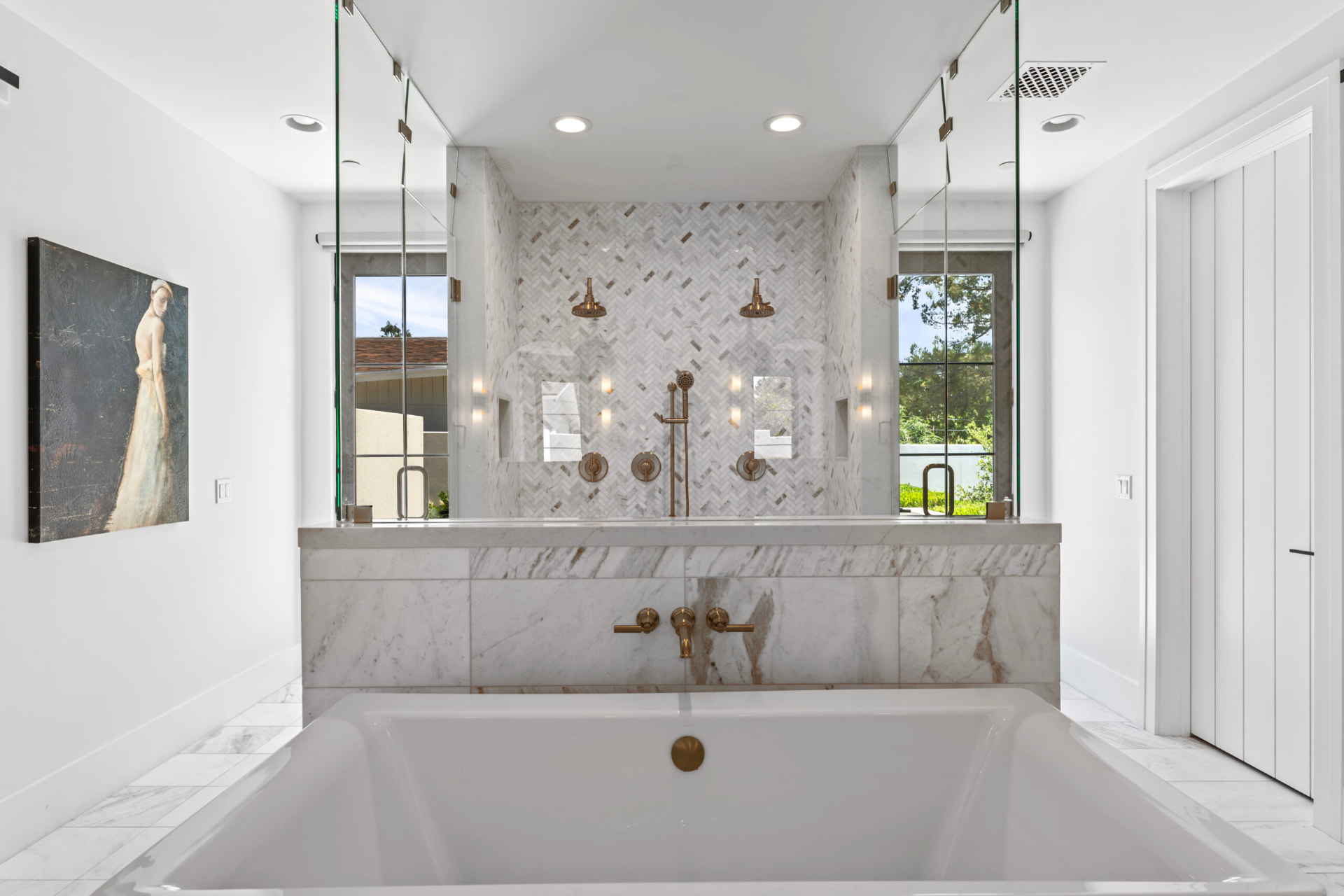 Bright symmetrical bathroom with a white soaking tub and brushed brass fixtures set against a white marble half-wall, behind which a glass-enclosed double shower shows chevron marble mosaic tile, dual brass showerheads and black-framed windows onto sunlit greenery
