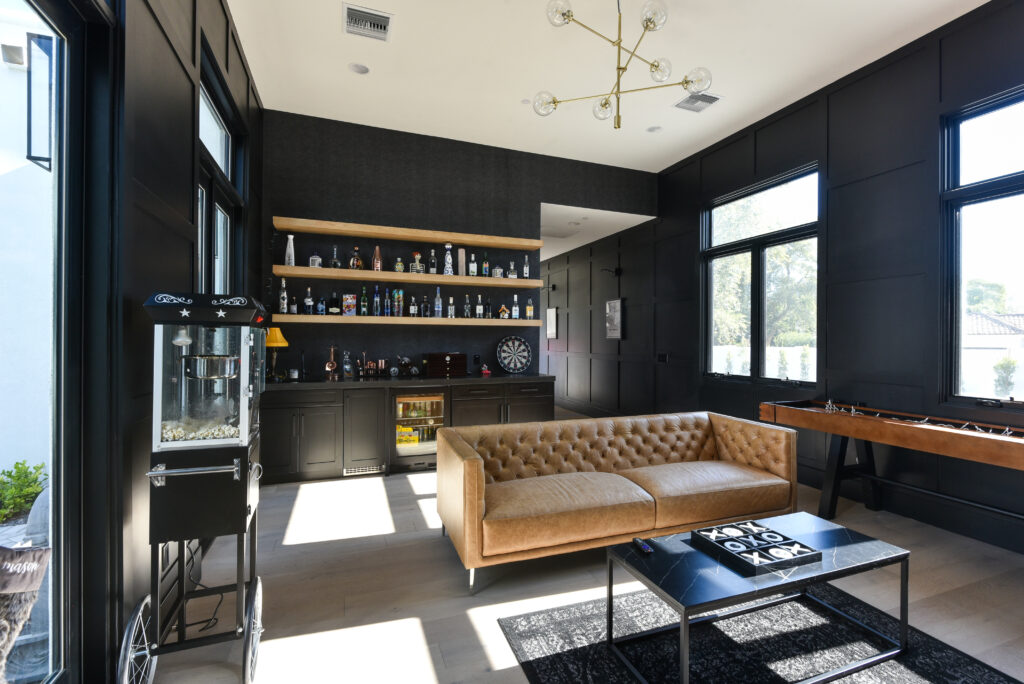 Game room with black paneled walls, floating wood shelves lined with bottles above a dark bar, a cognac tufted leather sofa, marble coffee table, shuffleboard table and a brass globe chandelier