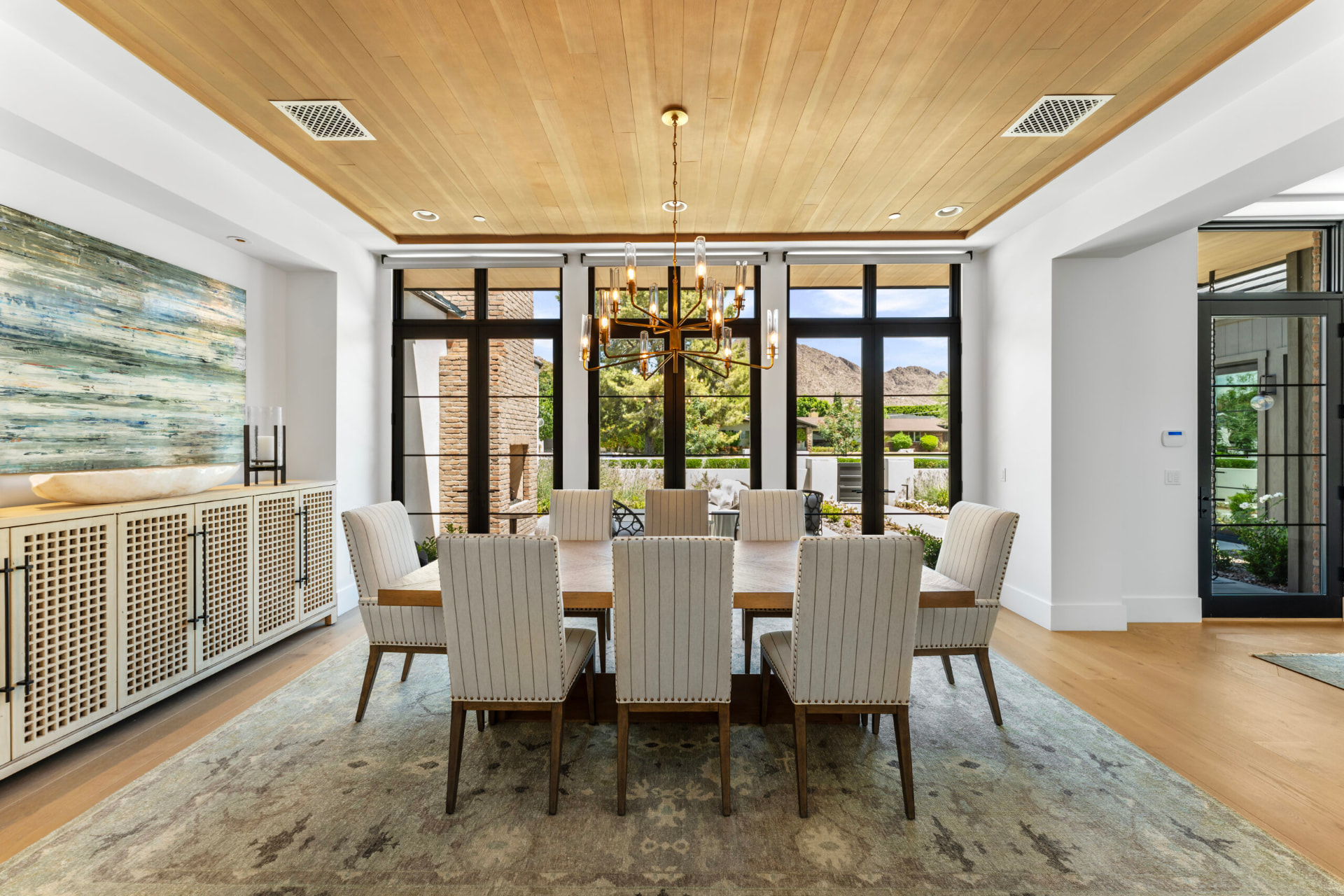 Dining room centred on a light-wood table with eight upholstered striped chairs on an antique-patterned rug, beneath a warm wood-paneled ceiling and brass chandelier, with black-framed French doors and windows looking out to a brick outdoor fireplace and desert hills
