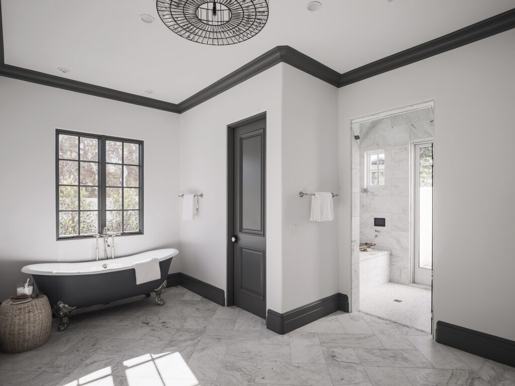 Primary bathroom with a matte black clawfoot soaking tub beneath a grid window, light gray marble tile floors, dark charcoal trim and doors, and a doorway into a marble walk-in shower