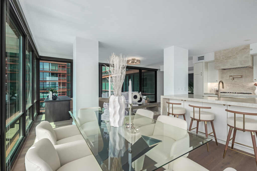Dining area with a glass table and cream chairs beside a kitchen island with barstools