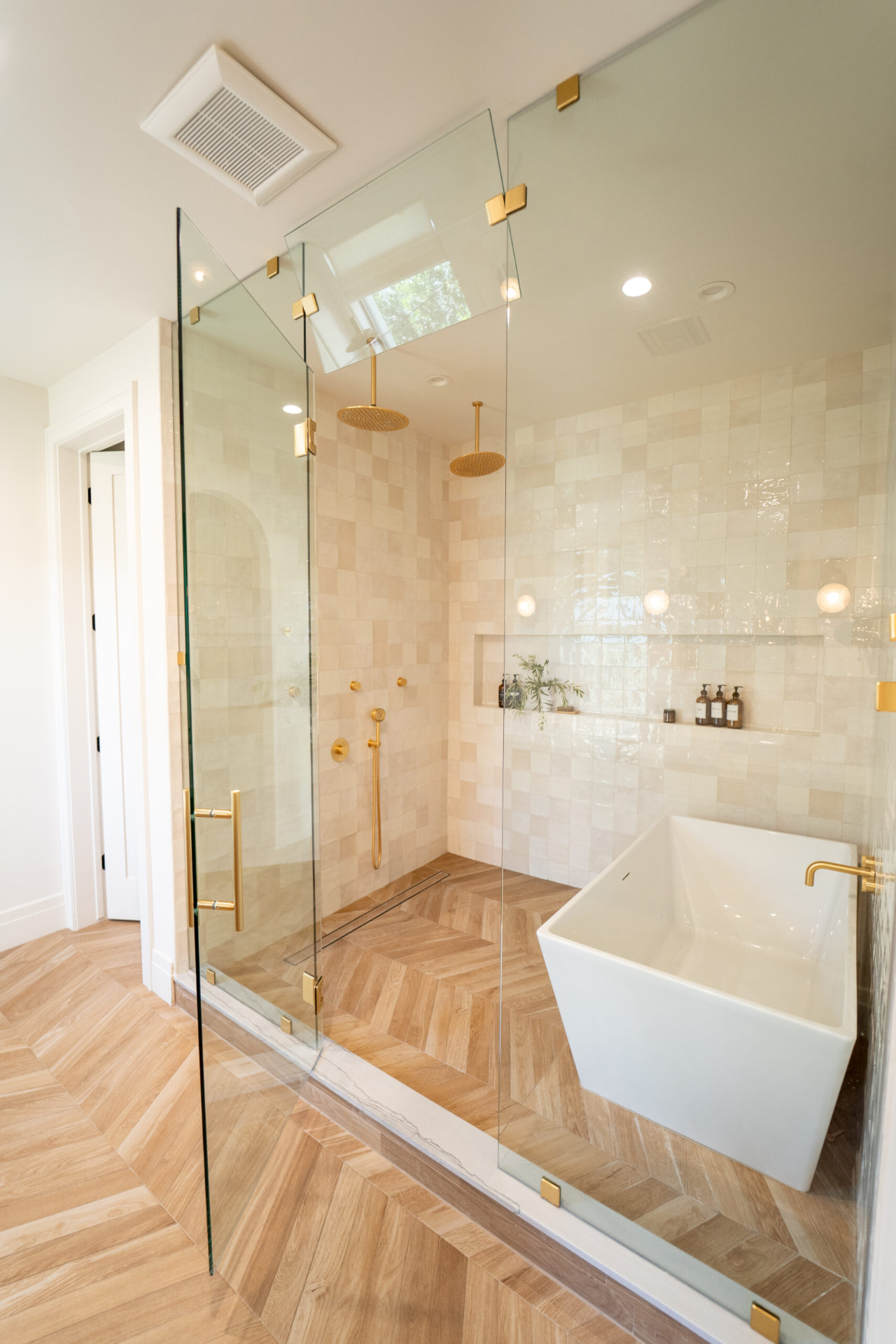 Bathroom with a freestanding white soaking tub behind clear glass with brass hardware, dual ceiling-mounted rain showerheads and light wood-look herringbone tile floors